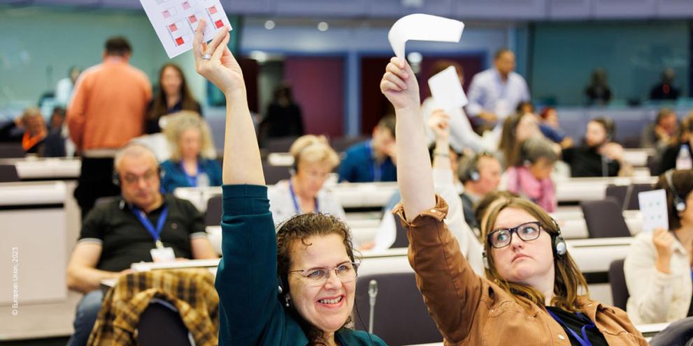 the image showcases 2 women sitting in a front row, raising up their hands with their vote.