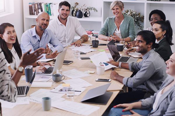 In this image, you can see a table where 8 business people from different age group and ethnicity are seated. They are discussing and they happy. They have laptops, papers and coffee mugs in front of them. 